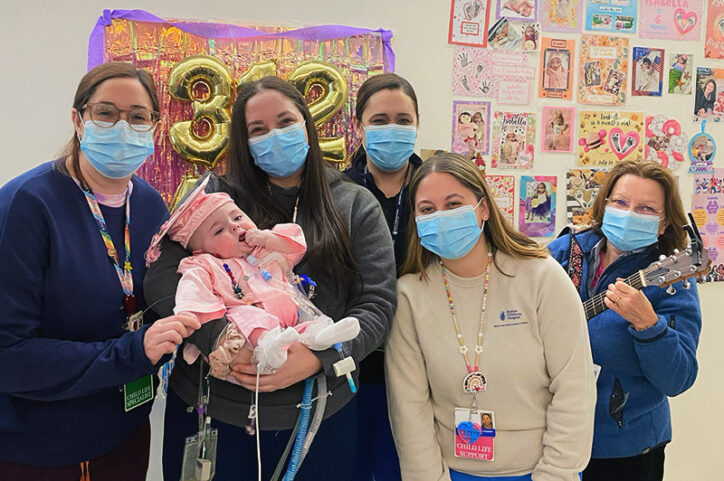 Care professionals surround a woman holding a baby with a tracheostomy and ventilator tubing during a milestone celebration. 