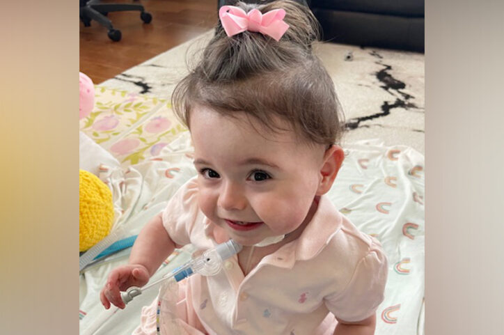 A toddler with a tracheostomy smiles while sitting on a blanket indoors.