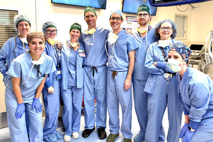 Nine medical professionals wearing scrubs, head coverings, and other protective clothing stand together in an operating room, smiling at the camera.