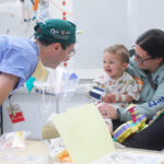 A doctor makes a toddler laugh while his mother holds him in a hospital bed.