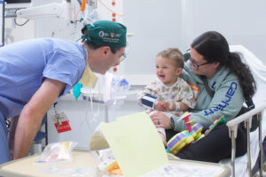 A doctor makes a toddler laugh while his mother holds him in a hospital bed.