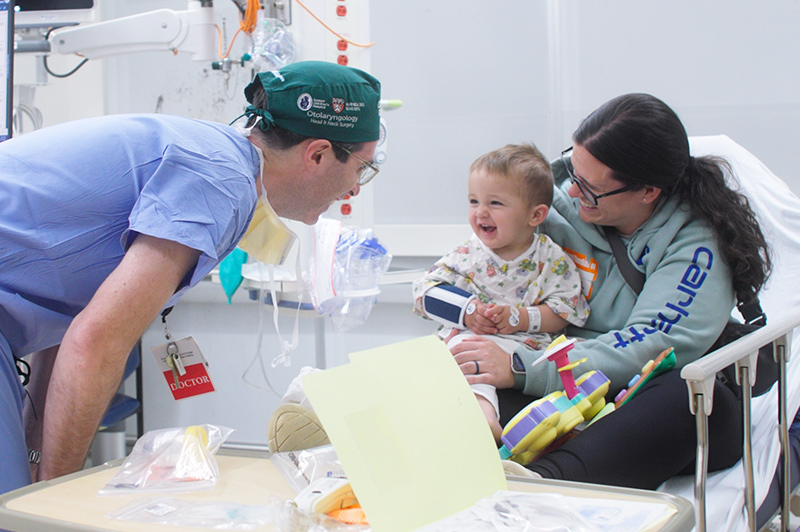 A doctor makes a toddler laugh while his mother holds him in a hospital bed.