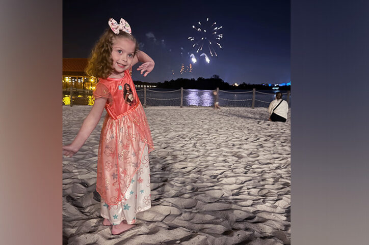 A young girl waves happily on a beach with firecrackers in the background. 