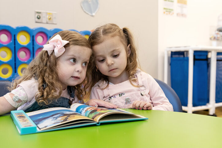 Two young girls lean into each other while reading a children's book. 