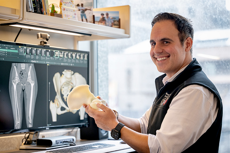 An engineer smiles while holding a 3D printed hip joint with an MRI image on a computer screen.
