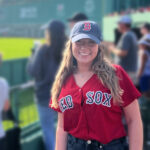 A young woman in her early 20s at Fenway Park wearing a Red Sox cap and jersey.