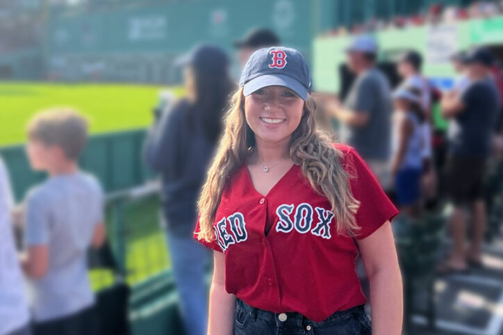 A young woman in her early 20s at Fenway Park wearing a Red Sox cap and jersey.
