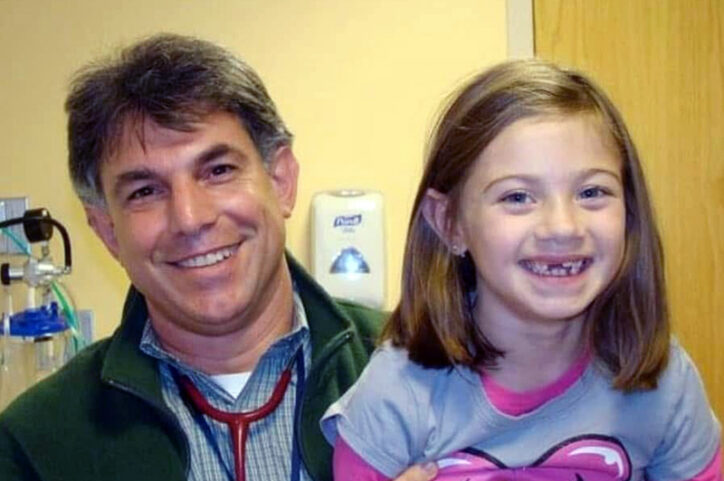 A smiling male doctor wearing a stethoscope sits beside a young girl during a medical visit.