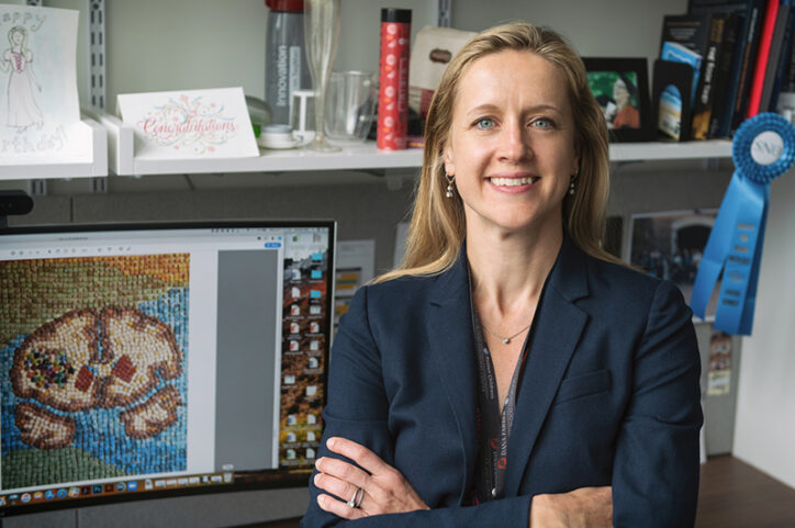 A woman in sits in an office with her arms crossed. A computer displays an image of a brain.