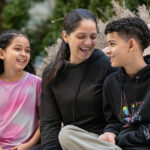 A woman, a girl, and a boy sit together outdoors, laughing and smiling at each other.