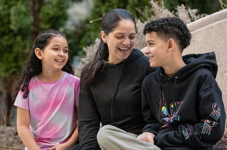A woman, a girl, and a boy sit together outdoors, laughing and smiling at each other.