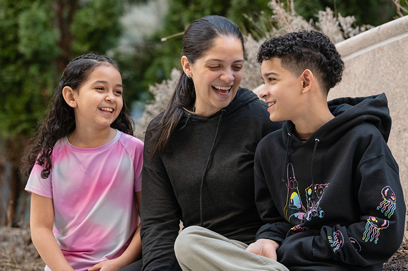 A woman, a girl, and a boy sit together outdoors, laughing and smiling at each other.