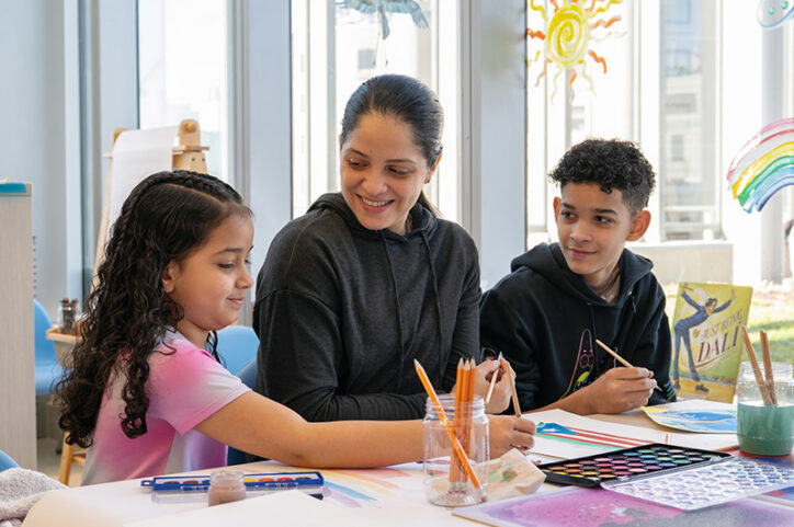A woman sits at a table between a young girl and a boy, smiling as they make art indoors.