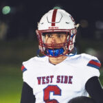 A teenage boy in a football helmet and uniform under stadium lights at night. His jersey reads: West Side