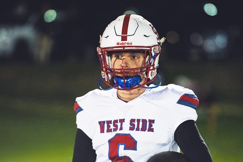 A teenage boy in a football helmet and uniform under stadium lights at night. His jersey reads: West Side