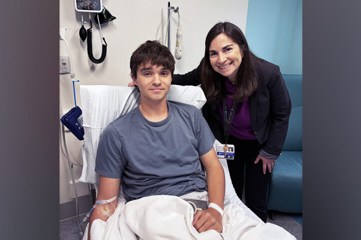A teenage boy with an IV in his arm sits in a hospital treatment chair while a woman in a blazer and hospital badge stands beside him.