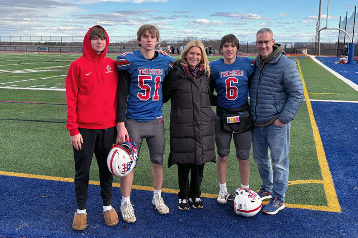 A teenage boy in a football uniform stands on a turf field with his parents and two brothers.