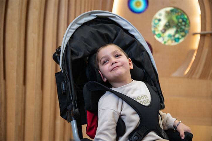 Child secured in a stroller with a padded harness, seated indoors in front of a wood wall with colorful circular decorations.