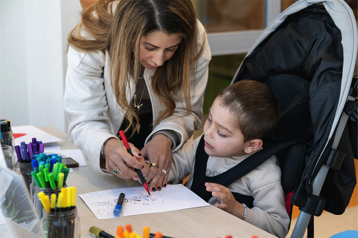 A woman helps a young child in a stroller color a piece of paper at a table with markers and drawing supplies.