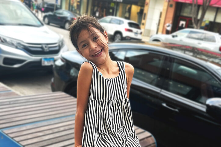 A 6-year-old girl in a striped dress smiles at the camera