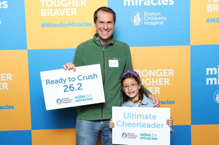 A 6-year-old girl and her father hold signs of encouragement in front of a backdrop that reads, "Miles for Miracles."