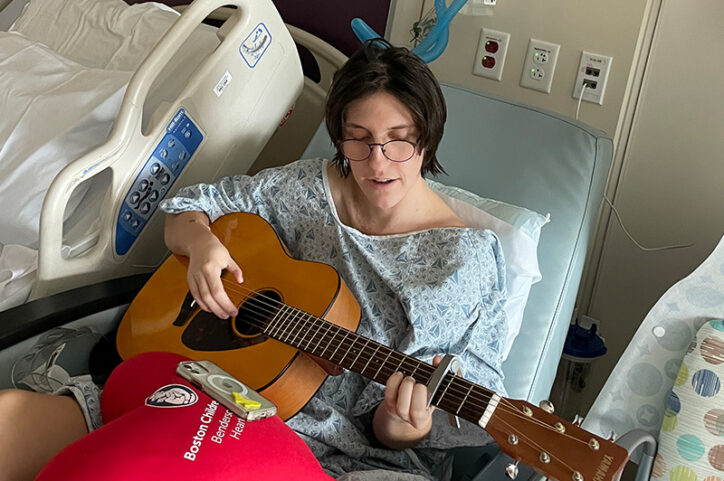 A young woman in glasses and a hospital gown plays guitar while in a hospital bed