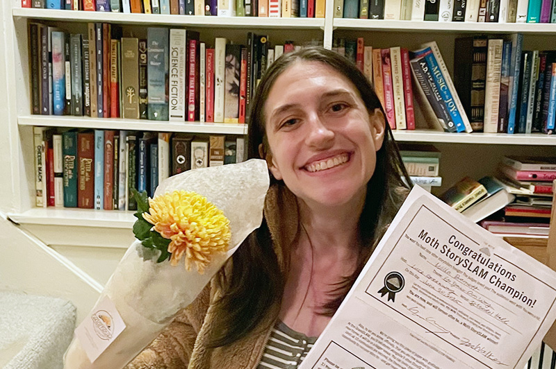 a smiling female young adult holds a bouquet of flowers and a certificate showing she won the Moth Story Slam
