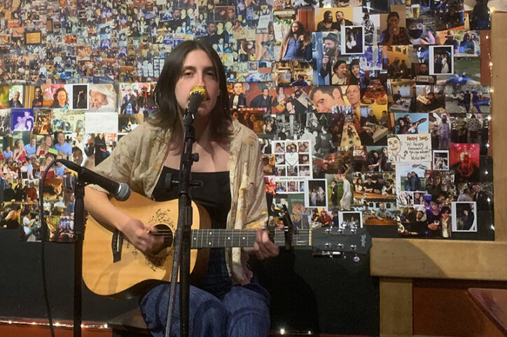 A young woman plays guitar and sings into a microphone in front of a wall of photographs