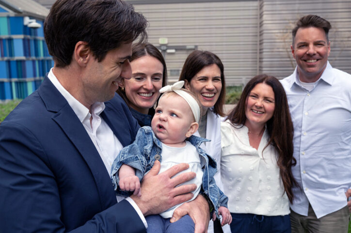 A smiling man holds a baby making a sour face, surrounded by four laughing adults in a garden setting.