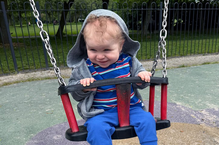 A young boy with a sweatshirt hoodie pulled over his head enjoys a ride on a swing.