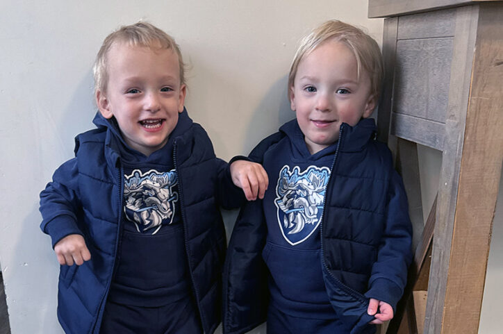 Twin toddler boys in sweatshirts and winter vests smiling.
