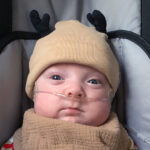 A baby in a reindeer cap and brown drool cloth sitting in a car seat.