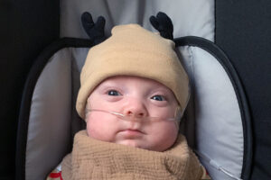 A baby in a reindeer cap and brown drool cloth sitting in a car seat.