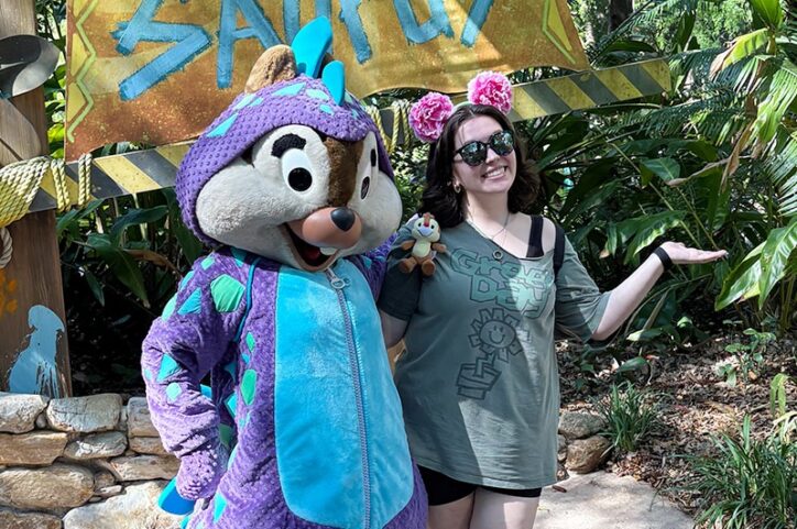 A young woman poses with a chipmunk mascot dressed as a dinosaur near a 'Chipmunk-O-Saurus' sign.