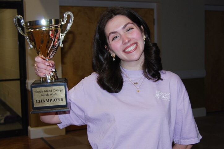 A young woman smiles while holding a trophy that reads: “Rhode Island College Greek Week Champions.” 