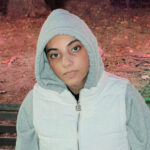 Young person in a gray hoodie and white vest sitting on a bench outside at night.