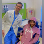 A male doctor in a white lab coat smiles with a young patient in a pink hoodie in a wheelchair in an exam room.