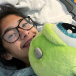 A smiling teen lies in her hospital bed holding a stuffed toy.