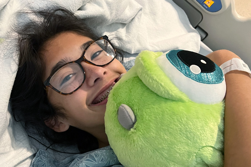 A smiling teen lies in her hospital bed holding a stuffed toy.