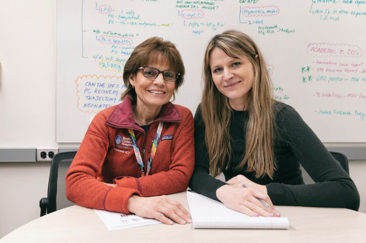 Two women smiling while seated at a table with a whiteboard behind them.