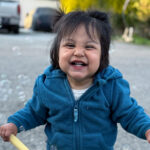 A toddler in a blue hoodie laughing while playing with bubbles.