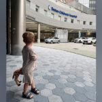 A young boy stands outside the entrance of Boston Children's holding a stuffed giraffe.