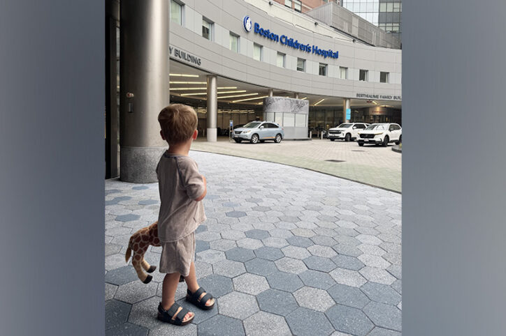 A young boy stands outside the entrance of Boston Children's holding a stuffed giraffe.