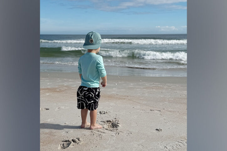 A young boy stands on the shore of a beach looking at breaking waves.